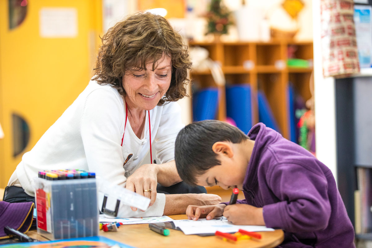 Teacher with brown curly hair shows young student painting at desk