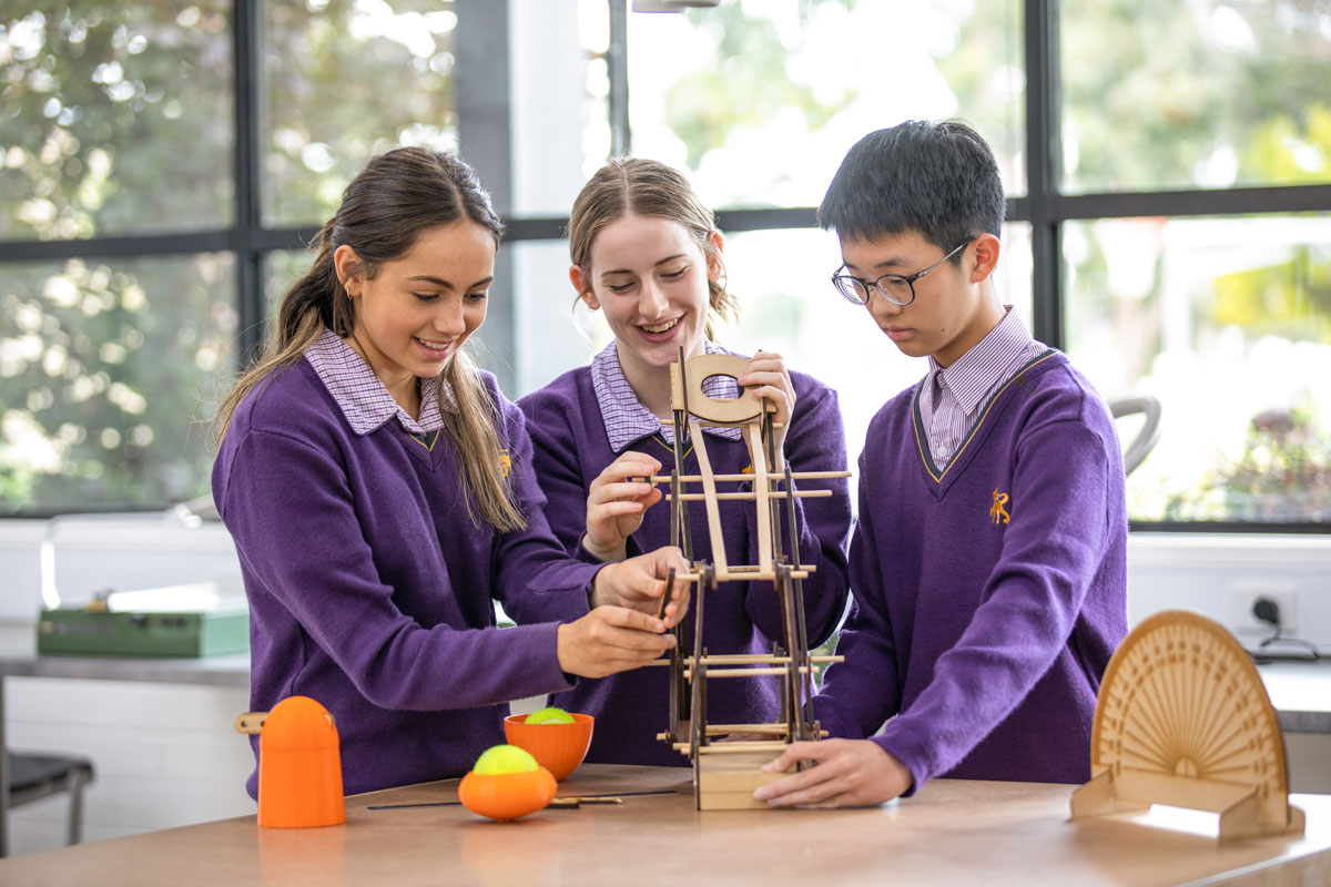 Three students construct a cardboard catapult on a table together