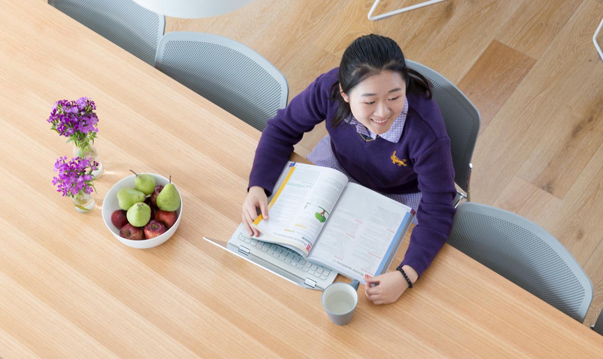 Student in school uniform studies at a desk with textbook and laptop